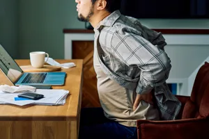 Man suffering back pain while working from home Man holding lower back in pain while sitting at a desk, struggling with poor posture during remote work