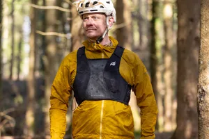 Bluegrass Venture Vest Mountain biker wearing a black Bluegrass Venture Vest over a yellow jacket, with a white helmet, standing in a forest.