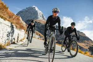 A Trio of Professional Cyclists, One of Them a Woman, Conquering the Mountains Together Front view photo of three elite cyclists - two men and one woman - dressed in full cycling gear as they tackle the winding mountain roads. Witness their unwavering focus and determination as they push themselves to the limit.