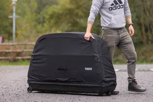 Thule RoundTrip MTB bike travel case Person pulling a large black Thule RoundTrip MTB bike travel case by its upper handle on a gravel surface.