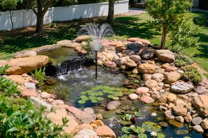 Solar-powered water fountain spraying water in a landscaped garden pond surrounded by rocks, water lilies and greenery. Solar-powered water fountain spraying water in a landscaped garden pond surrounded by rocks, water lilies and greenery.