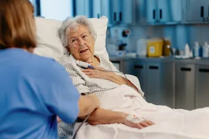 Senior patient feeling pain on chest, heart attack symptoms for elderly woman. Nurse checking on senior woman in emergency room. Healthcare and emotional support for elderly patient. An older woman clutching her chest while lying in a hospital bed