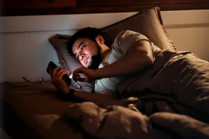 Man suffering insomnia in bed checking phone and laptop A man lying in his bed scrolling on his phone