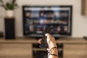 A woman is holding a remote control of a smart TV in his hand. In the background you can see the television screen with streaming entertainment apps for video on demand. Close up of a person holding a control remote with a television screen on the background. A woman holding a remote control switches programs on the TV in the background.