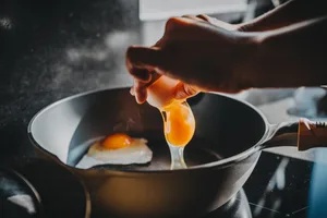 Close-up of female hand cracking an egg into a sizzling hot frying pan as she prepares a healthy breakfast in kitchen in the morning. Clean eating and healthy eating habits with natural ingredients. Wellness lifestyle and mindful meal preparation Close-up of female hand cracking an egg into a sizzling hot frying pan