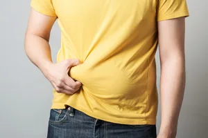 biggest-factor-where-you-gain-fat Close up of overweight man's hand holding his belly over gray background.