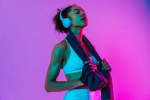 BPM A woman wearing activewear and headphones is standing in front of a pink gradient background with her eyes closed