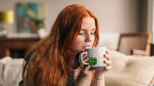 Enjoying a warm cup of Pukka herbal tea at home in a comfortable, relaxed setting. A person with long red hair holding and drinking from a Pukka herbal tea mug while sitting on a sofa in a cozy living room.