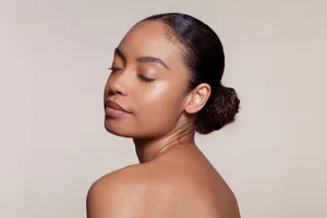 Easy ways to boost collagen naturally (1) A woman with glowing skin stands with her eyes closed in front of a beige background