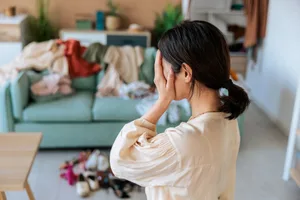 Mental health benefits of cleaning A woman stands in a messy living room, covering her face with her hands