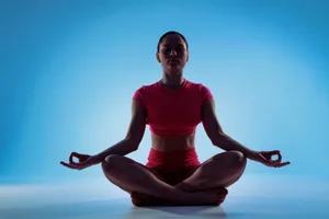A woman wearing red activewear mediates in a seated position against a blue background A woman wearing red activewear mediates in a seated position against a blue background