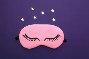 A pink sleep mask next to a cluster of stars on a purple background A pink sleep mask next to a cluster of stars on a purple background