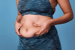 Cortisol belly A woman wearing activewear is squeezing her stomach in front of a blue background