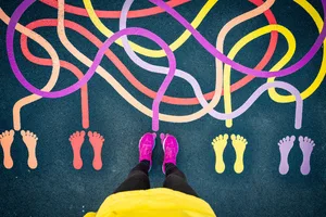 How far do you really need to walk to be healthy An overhead shot of a person wearing bright pink shoes standing in a playground is shown. On the ground, there are painted colourful lines leading to different sets of footprints in orange, yellow and purple.