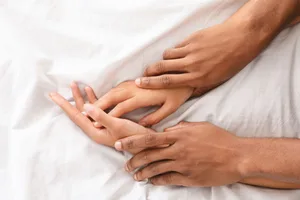 Signs your partner is faking it during sex A close-up of a man gently holding a woman's wrists on a white bed sheet