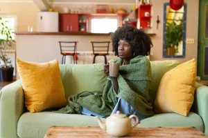 Main image (3) A woman is huddled on her sofa, snuggled into a large blanket around her shoulders as she holds a mug of a hot tea, trying to warm up and feel better while suffering from a cold or flu