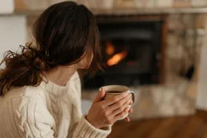 Tricks for heating your home more efficiently A woman holding a hot drink is sat by a fireplace