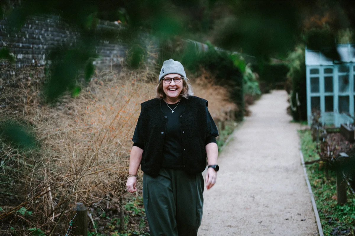 Native_7RA6115-02 Kate, a middle-aged woman wearing a grey hat and smiling as she walks towards the camera in her garden