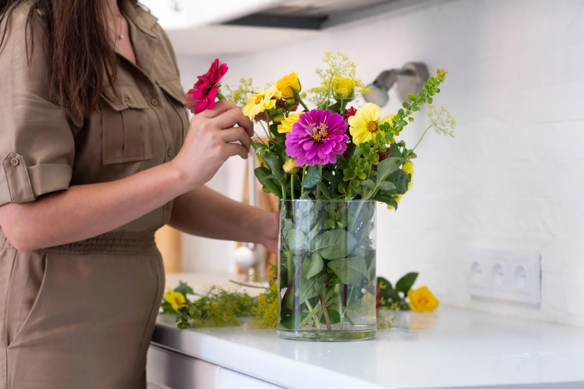 A woman arranging a colourful bouquet in a decorative vase on a kitchen counter.